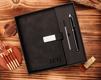 May include: A black leather journal with the initials "MH" engraved on the cover, presented in a gift box with a pen and a pen refill. The journal has a silver clasp. A golf tee holder and other accessories are also visible.