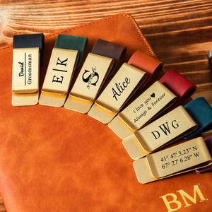 Puede incluir: Una colección de pinzas para billetes personalizadas con varios nombres, iniciales y mensajes, dispuestas sobre un cuaderno de cuero. Las pinzas tienen detalles de cuero de diferentes colores y son de metal dorado. Un bolígrafo negro descansa junto al cuaderno.