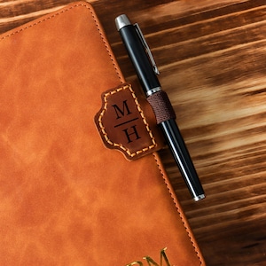 May include: A close-up of a brown leather journal with a pen resting on top. The journal has a stitched leather patch with the initials "M H". The pen is black with a silver tip and a textured brown grip. The journal is on a wooden surface.