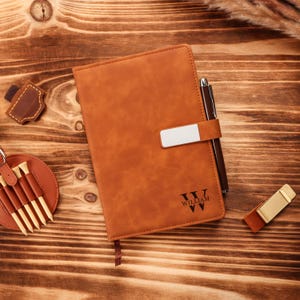 May include: A brown leather journal with a silver clasp and a pen. The journal is personalized with the name "WILLIAM". A golf tee holder and other leather accessories are also visible.