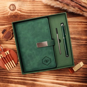 May include: A green leather journal and pen set in a gift box. The journal has a silver clasp and the initials "JWF" in a hexagon. The pen is green with silver accents. The set is on a wooden surface.