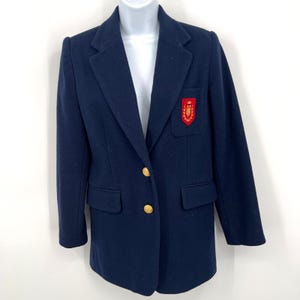 May include: A navy blue blazer with gold buttons and a red embroidered crest on the chest. The blazer has a classic tailored design with a notched lapel and two front pockets. The blazer is displayed on a white mannequin.