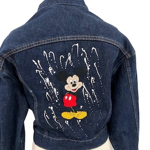 May include: Dark blue denim jacket featuring a Mickey Mouse design on the back. The design includes a black Mickey Mouse with a red shirt, yellow shoes, and white paint splatter accents. The jacket has a classic collar and button closure.
