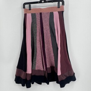 May include: A patchwork skirt with a variety of colors, including pink, gray, and burgundy. The skirt is made of different fabrics and textures, with a black hemline and a pink waistband. The skirt is hanging on a black hanger.