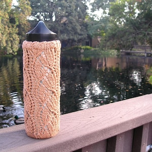 May include: A knitted bottle cozy in a light peach color with a delicate lace pattern. The cozy is on a wooden railing overlooking a pond with ducks swimming in the water.
