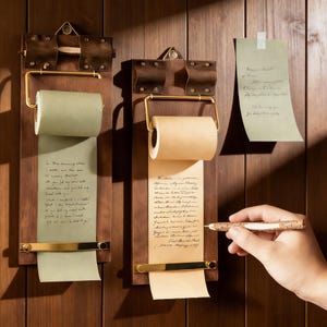 Puede incluir: Dos portarrollos de notas de madera con detalles de latón están montados en una pared de madera. Uno contiene un rollo de papel verde, el otro un rollo de papel marrón. Una mano sostiene un bolígrafo, escribiendo en una nota suelta.