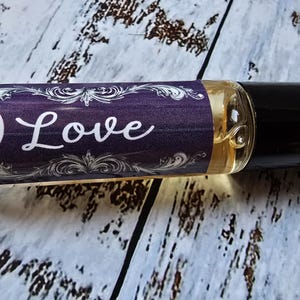 May include: A clear glass roll-on bottle with a black cap. The label is purple with white ornate designs and the word "Love" in white script. The bottle contains a golden liquid and is set against a light wood grain background.