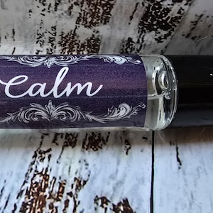 May include: A clear glass roll-on bottle with a black cap. The label is purple with white text that reads "Calm" and decorative scrollwork. A small logo is visible on the left side of the label.