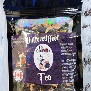 May include: A black resealable pouch with a clear window showing a blend of black tea, butterscotch chips, calendula flowers, and white chocolate. The pouch is labelled "Buttered Beer Tea" and "The Cauldron". The text on the pouch also includes instructions for brewing the tea. The pouch has a "Proudly Made in Canada" sticker.