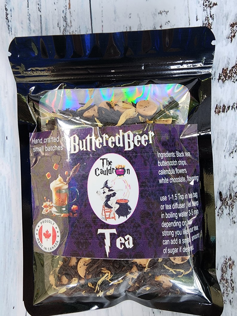 May include: A black resealable pouch with a clear window showing a blend of black tea, butterscotch chips, calendula flowers, and white chocolate. The pouch is labelled "Buttered Beer Tea" and "The Cauldron". The text on the pouch also includes instructions for brewing the tea. The pouch has a "Proudly Made in Canada" sticker.