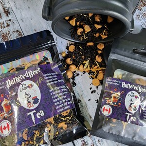 May include: Two black zippered pouches with a purple and black label that reads "Buttered Beer The Cauldr Tea" and a Canadian flag. The pouches contain loose leaf tea and are sitting on a white wooden surface.
