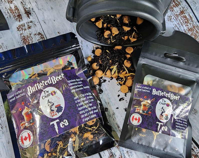 May include: Two black zippered pouches with a purple and black label that reads "Buttered Beer The Cauldr Tea" and a Canadian flag. The pouches contain loose leaf tea and are sitting on a white wooden surface.