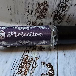 May include: A clear glass roll-on bottle with a black cap. The label is purple with white script that reads "Protection". The label also features a small illustration and decorative flourishes. The bottle is on a white and grey surface.