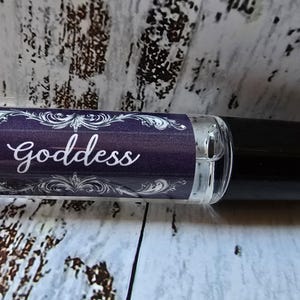May include: A clear glass roll-on bottle with a black cap. The label is dark purple with white ornate designs and the word "Goddess" in elegant script. A small logo is visible on the left side of the label.