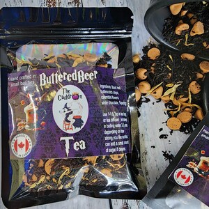 May include: A black pouch with a purple and white label that says "Buttered Beer" and "The Cauldron Tea". The label has a witch illustration and text about the ingredients and how to brew the tea. The pouch is open and shows the tea inside, which includes black tea, butterscotch chips, calendula flowers, and white chocolate.