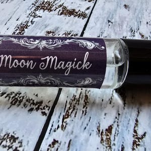 May include: A clear glass roll-on bottle with a black cap. The label is dark purple with white ornate designs and the words "Moon Magick". The bottle is on a light wood-grain surface.