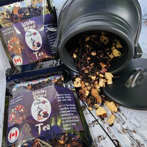 May include: Two black and purple tea bags with the text "Witches Harvest The Cauldron Tea" and a black cauldron with a red and orange tea blend spilling out. The tea blend includes black tea, cocoa nibs, cinnamon, apple, cloves, and vanilla.