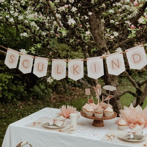 May include: A white tablecloth with pink and gold confetti and two place settings.  A wooden cake stand with cupcakes decorated with pink frosting and toppers.  A banner with the word "SULKIND" in pink and gold letters hangs above the table.