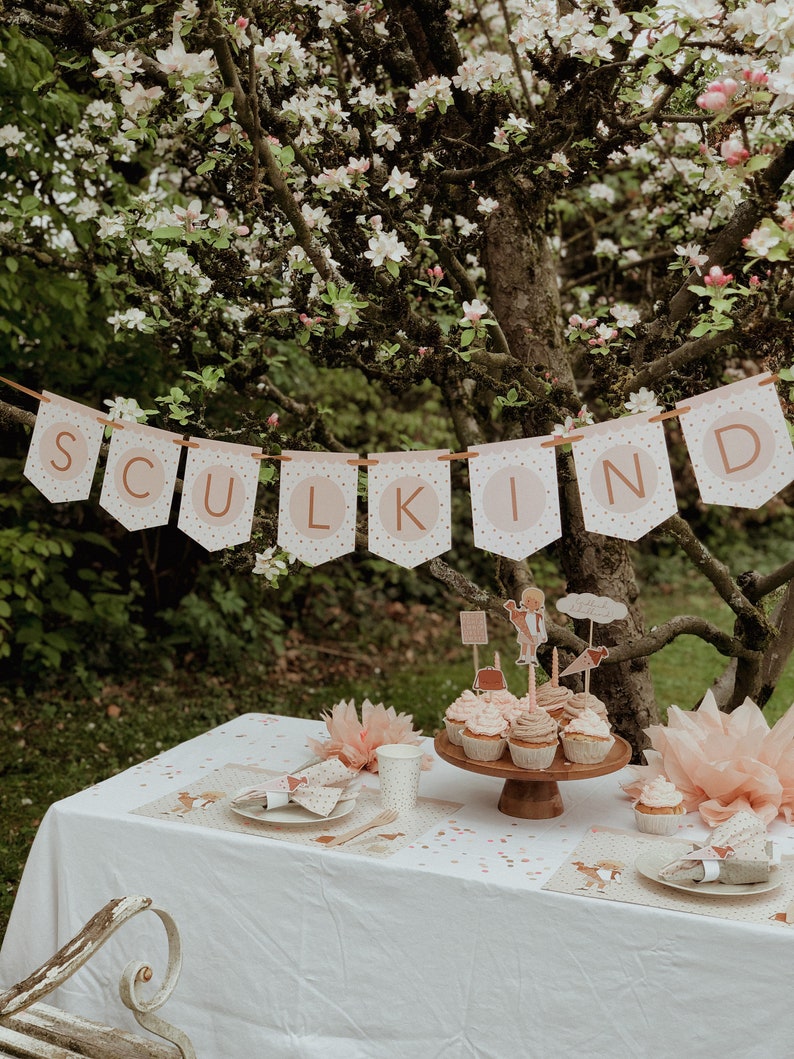 May include: A white tablecloth with pink and gold confetti and two place settings.  A wooden cake stand with cupcakes decorated with pink frosting and toppers.  A banner with the word "SULKIND" in pink and gold letters hangs above the table.