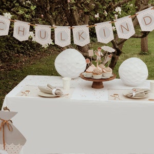Könnte beinhalten: Eine weiße Tischdecke mit einem Banner, auf dem "SCHULKIND" in braunen Buchstaben steht. Auf einem hölzernen Kuchenständer stehen Cupcakes mit rosa Zuckerguss und Toppern. Auf dem Tisch stehen zwei weiße Papierlaternen und ein weißer Papierbecher. Links auf dem Tisch befindet sich eine graue Papiertüte mit einem braunen Band.