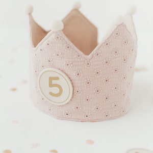 Könnte beinhalten: Eine rosa Stoffkrone mit weißen Bommeln und einem goldenen Kreis mit der Zahl "5" darauf. Die Krone liegt auf einer weißen Oberfläche mit goldenen und rosa Konfetti.