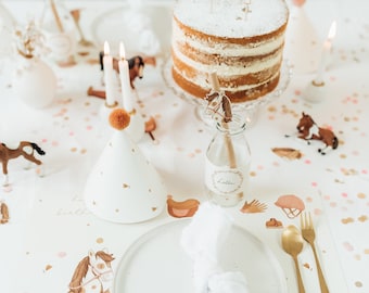 May include: A white table setting with a horse-themed party decor. The table is covered in a white tablecloth with a brown and pink horse print. There is a white cake with a horse topper, a white party hat, a glass bottle with a horse straw, and gold cutlery. The text "happy birthday!" is on the cake topper.