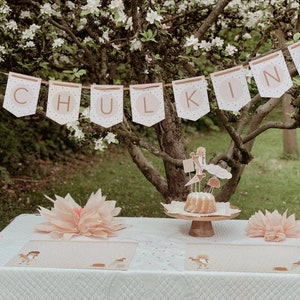 May include: A white tablecloth with two place settings, each with a pink paper doily and a small brown and white patterned placemat. A cake with white frosting and a topper featuring a girl and a cloud sits on a wooden cake stand in the centre of the table. A banner with the word "SCHULKIND" hangs from a tree branch above the table.