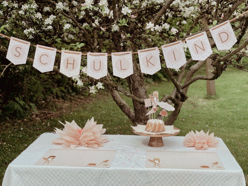 May include: A white tablecloth with two place settings, each with a pink paper doily and a small brown and white patterned placemat. A cake with white frosting and a topper featuring a girl and a cloud sits on a wooden cake stand in the centre of the table. A banner with the word "SCHULKIND" hangs from a tree branch above the table.