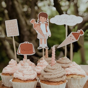 May include: A set of cupcakes decorated with pink and brown frosting. The cupcakes are topped with various school-themed decorations, including a girl holding a school cone, a backpack, and a cone with a smiley face. The text "endlich schulkind!" is visible on one of the decorations.