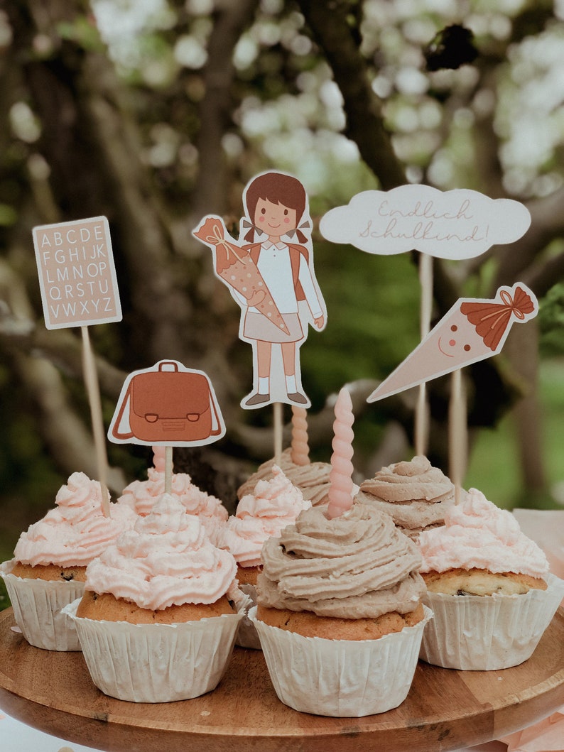 May include: A set of cupcakes decorated with pink and brown frosting. The cupcakes are topped with various school-themed decorations, including a girl holding a school cone, a backpack, and a cone with a smiley face. The text "endlich schulkind!" is visible on one of the decorations.