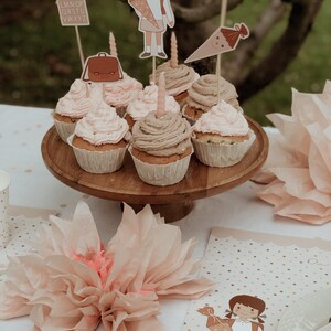 May include: A table setting for a first day of school party with cupcakes decorated with school-themed toppers. The cupcakes are on a wooden cake stand. There are also tissue paper decorations and a place card with the text "Endlich Schulkind!" on the table.