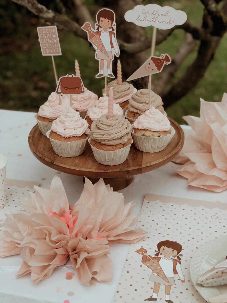 May include: A table setting for a first day of school party with cupcakes decorated with school-themed toppers. The cupcakes are on a wooden cake stand. There are also tissue paper decorations and a place card with the text "Endlich Schulkind!" on the table.