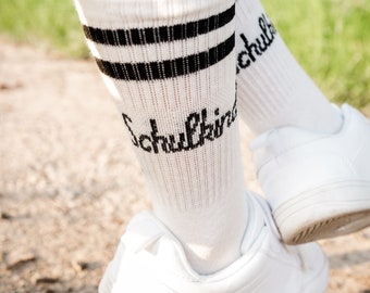 May include: White knee-high socks with black stripes and the word "Schulkind" written in black. The socks are worn with white trainers.