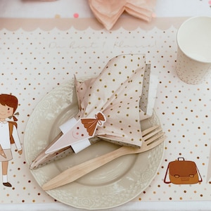 May include: A pink and brown polka dot placemat with a brown school bag, a brown and white school cone, and a girl holding a school cone. The place setting includes a white plate, a wooden fork, and a pink and brown polka dot napkin.