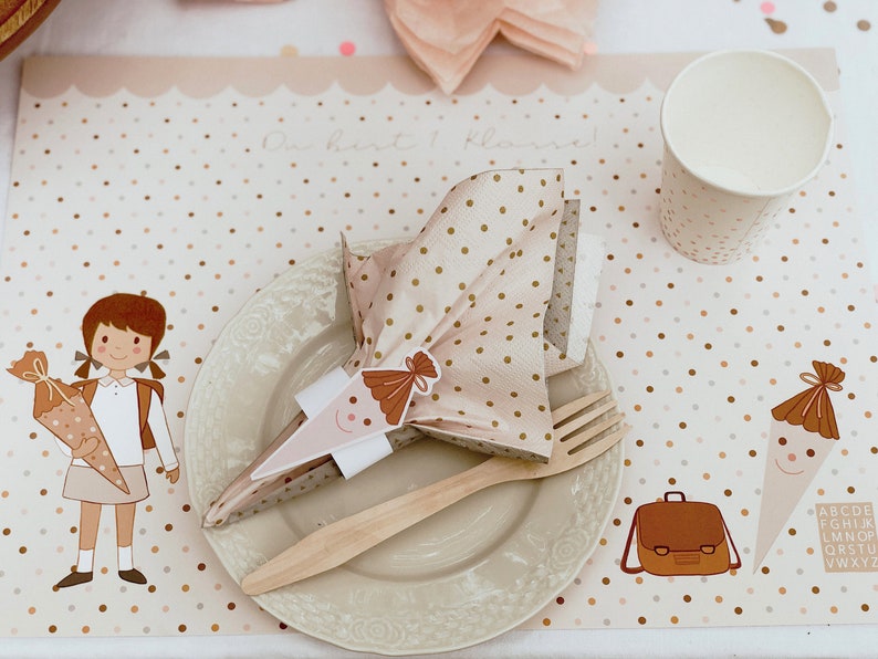 May include: A pink and brown polka dot placemat with a brown school bag, a brown and white school cone, and a girl holding a school cone. The place setting includes a white plate, a wooden fork, and a pink and brown polka dot napkin.