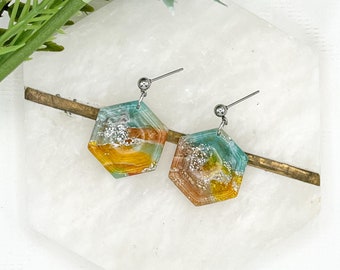 May include: A pair of hexagonal resin earrings with a teal, orange, and gold color scheme. The earrings have a silver post and a silver bead at the top.