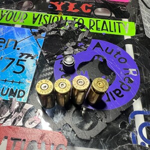 May include: Four gold-colored bullet casings with the word "FEDERAL" printed on them. The casings are on a black surface with various stickers and a purple sticker with the text "Auto Repair" on it.