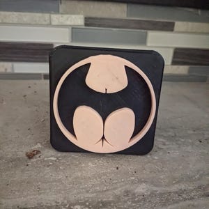 May include: A black square object with a light tan Batman logo. The logo is a stylized bat symbol within a circle. The object is sitting on a gray surface.