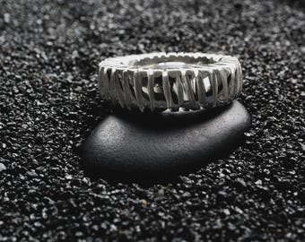 May include: A silver ring with a distinctive, open design of vertical bars. The ring sits on a smooth, black stone, against a background of dark, granular material. This piece of jewellery is a modern and minimalist accessory.