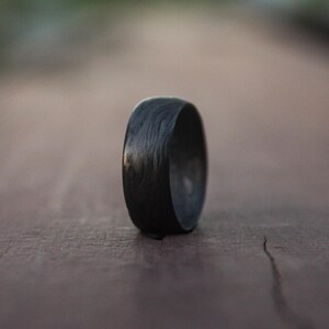 以下が含まれることがあります： 木目模様のユニークな黒いカーボンファイバーリング。リングは木製の表面の上に置かれています。