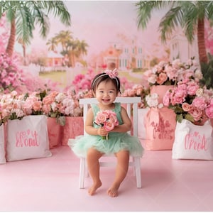 May include: A young girl in a mint green tulle dress sits on a white chair holding a bouquet of pink roses. She is wearing a headband with a pink flower. The background is a pink and green floral backdrop with a pink and white floral arrangement on either side of the chair.