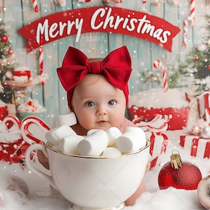 Könnte beinhalten: Ein Baby mit einem roten Schleifenband sitzt in einer weißen Teetasse, die mit weißen Marshmallows gefüllt ist. Das Baby schaut in die Kamera. Der Hintergrund ist eine Weihnachtskulisse mit einem roten Banner, auf dem "Frohe Weihnachten" steht.