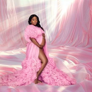 May include: A pregnant woman in a pink ruffled gown poses in a studio setting with a pink backdrop. The gown has a long train and a thigh-high slit. The woman is looking down at her stomach.