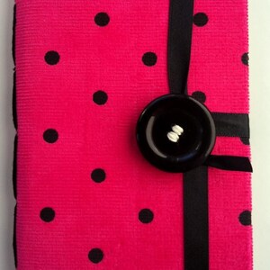 May include: A pink notebook with black polka dots and a black ribbon closure. A large black button is attached to the ribbon.