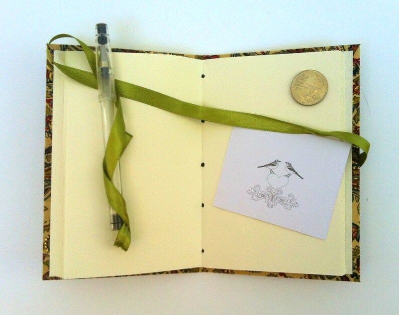 Puede incluir: Diario abierto con p&aacute;ginas color crema, una cinta verde y un bol&iacute;grafo transparente. La cubierta tiene un dise&ntilde;o estampado en oro, rojo y verde. Una peque&ntilde;a tarjeta con una ilustraci&oacute;n de p&aacute;jaro y una moneda est&aacute;n dentro.