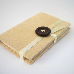 May include: A beige corduroy notebook with a white ribbon tied around it and a brown button in the center of the ribbon.