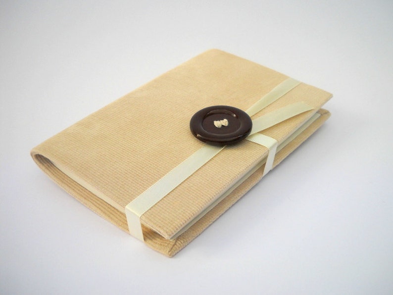 May include: A beige corduroy notebook with a white ribbon tied around it and a brown button in the center of the ribbon.