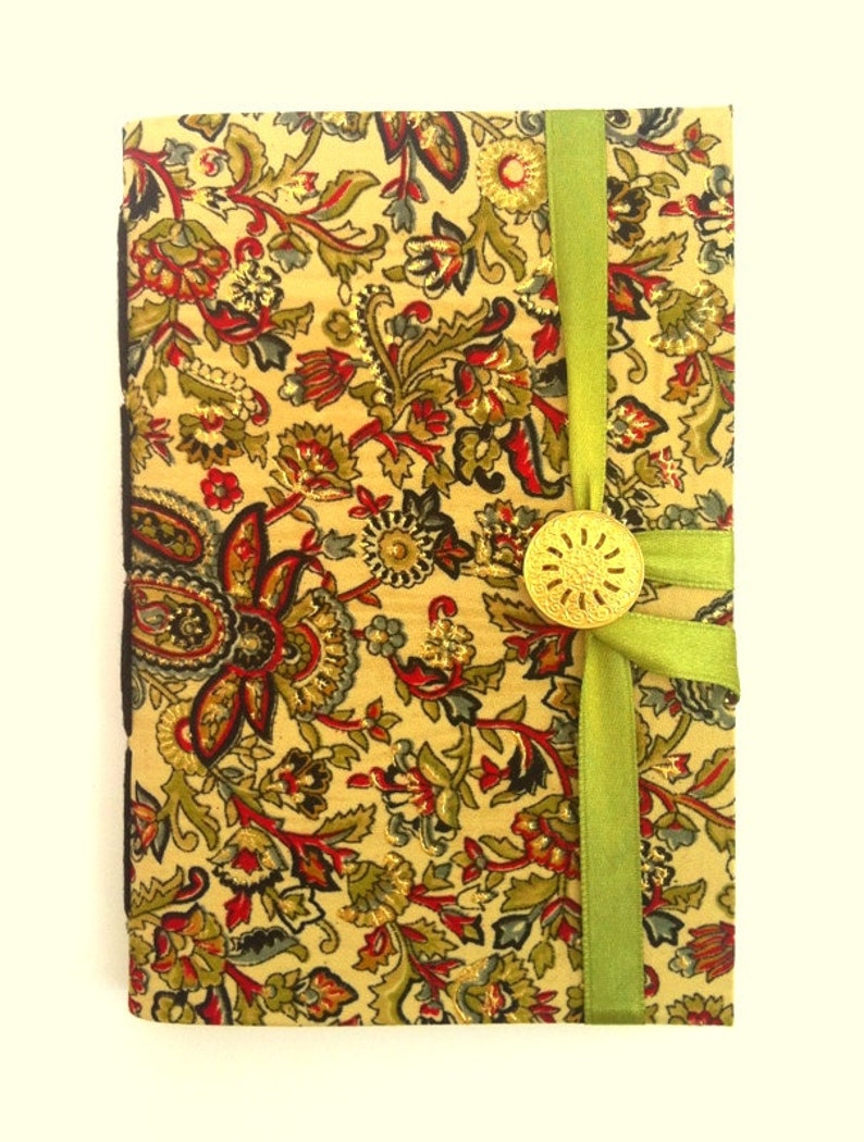 Puede incluir: Un diario cubierto de tela con un estampado floral en rojo, verde y dorado sobre un fondo crema. Una cinta verde envuelve el diario, asegurada con un bot&oacute;n dorado. Los bordes del diario son negros.