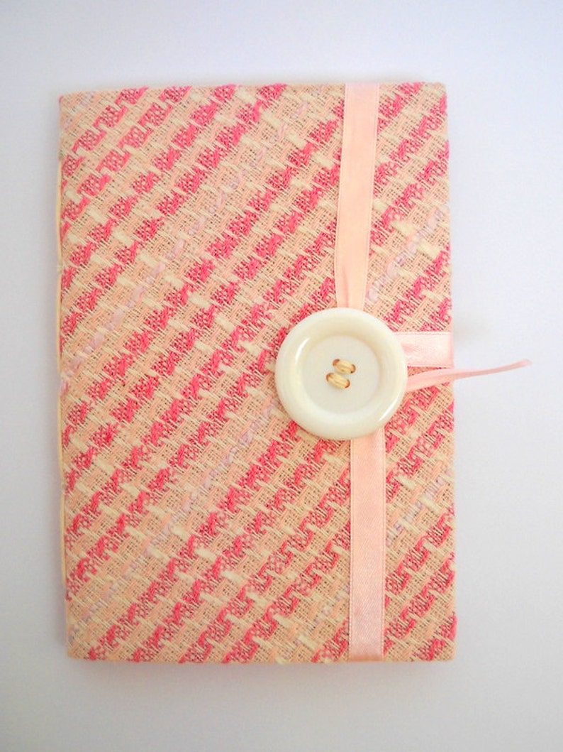 Puede incluir: Un cuaderno de tela con estampado rosa y blanco, con una cinta rosa y un bot&oacute;n blanco.