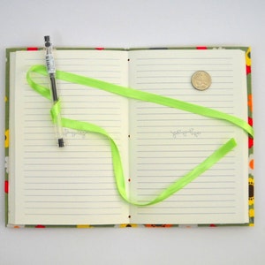 May include: A green ribbon is tied around a closed notebook with a floral pattern on the cover. A pen and a coin are on the open pages.